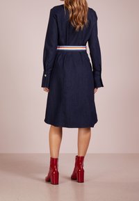 Tummansininen farkkumekko, jossa on istuva vyötärö ja pitkät hihat. Mekossa on värikäs raidallinen vyö, ja se on yhdistetty kiiltäviin punaisiin nilkkureihin.