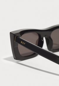 Black sunglasses with angular design and brown lenses. Features gold “RSF” lettering on the matte black temple. Smooth, glossy texture.