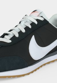 Ασπρόμαυρο αθλητικό παπούτσι Nike με λευκά κορδόνια, μεγάλο λευκό σήμα στη πλευρά και καουτσούκ σόλα με υφή σε απλό φόντο.