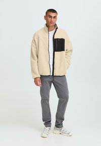 Junger Mann steht in einem beigen Fleece-Jacke mit schwarzer Brusttasche, weißem Hemd, grauen Jeans und weißen Sneakern vor einem einfarbigen Hintergrund.