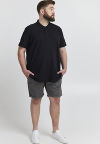 Camisa polo negra, pantalones cortos grises y zapatillas blancas. La polo tiene un cuello y botones, mientras que los pantalones cortos tienen un dobladillo enrollado.