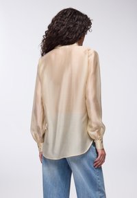 Blusa beige chiaro, semi-trasparente, con maniche lunghe, polsini con bottoni e orlo curvo. I jeans in denim abbinati hanno un aspetto sbiadito.