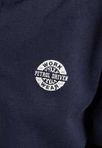 Tessuto blu navy con un logo circolare bianco che reca la scritta "WORK WEAR PETROL DRIVEN" e un motivo di battistrada al centro.
