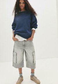 Felpa navy oversize con orlo bianco a strati, abbinata a pantaloni cargo grigi chiari con grandi tasche laterali. Sneakers beige completano il look.