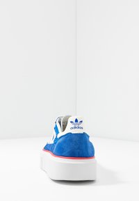 Blue suede sneaker with a white rubber sole, featuring three white stripes, a red accent at the base, and an Adidas logo on the heel.