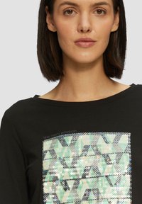Schwarzes Langarmshirt mit geometrischem Paillettenmuster in den Farbtönen Grün, Blau und Silber auf der Vorderseite. Weiches Textilmaterial.