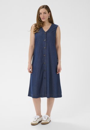 Femme portant une robe midi bleue sans manches à boutons et des baskets blanches, debout devant un fond clair uni.