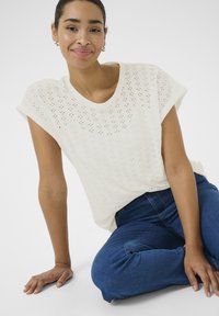T-shirt en tricot perforé blanc avec des manches courtes retournées, associé à un jean en denim bleu. Le haut présente un col rond et une coupe ample.