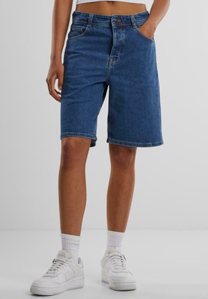 Jeanshorts i mørkeblå med en avslappet passform, fem lommer og rullet kant. Matchet med hvite sneakers og ankelsokker.