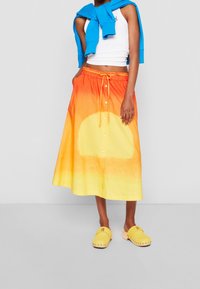 Mehrfarbiger Rock mit Farbverlauf von Orange zu Gelb, Kordelzugtaillenbund, Knopfdetails, kombiniert mit gelben Slippers und einem blauen Pullover.