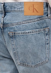 Τζιν παντελόνι από ανοιχτό μπλε τζην με πίσω τσέπη που έχει δερμάτινο ταμπελάκι σε ταμπά χρώμα, με το λογότυπο της μάρκας "cK" ραμμένο πάνω από την τσέπη.