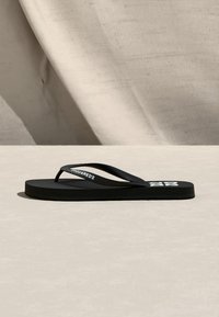 Sandale noire de type tongs avec "DSQUARED2" sur la bride et le logo "D2" sur la semelle, exposée sur une surface texturée beige avec un fond assorti.