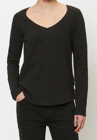 Person wearing a black long-sleeve V-neck shirt and black pants, with one hand in pocket against plain light background.