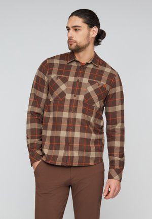 Uomo con capelli scuri legati dietro, indossa una camicia a quadri marrone e beige e pantaloni marroni, guarda a sinistra con una mano in tasca.