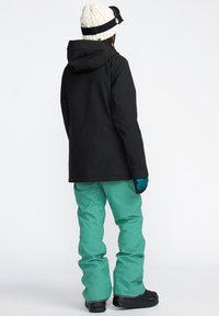 Schwarze wasserdichte Jacke und türkisfarbene Skihose, mit einer gestrickten weißen Mütze und Handschuhen. Glatte Textur, verstellbare Kapuze und verstärkte Nähte.