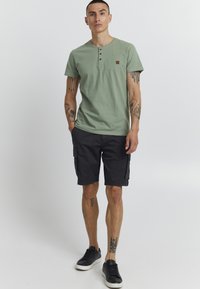 T-shirt vert à col rond avec deux boutons et un petit badge logo marron, associé à un short cargo noir et des baskets noires.