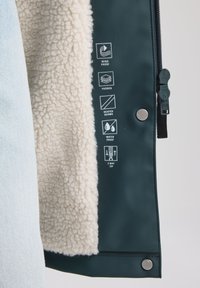 Veste imperméable avec un tissu extérieur vert foncé, doublure en polaire beige et divers symboles d'entretien. Comprend une fermeture éclair à double sens et des boutons pression.