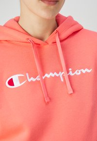 Felpa corallo con cappuccio e lacci, con un logo "Champion" bianco ricamato e un accento rosso nella "C" del logo. Tessuto morbido e texturizzato.