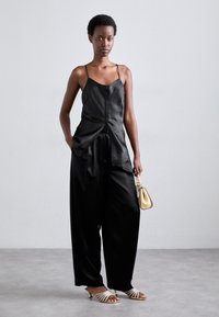 Camisole en satin noir avec des détails de boutons, associé à un pantalon noir large. Accessoirisé avec un petit sac à main doré et des talons à lanières.