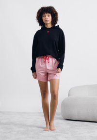 Zwarte hoodie met een rood logo, gecombineerd met roze shorts met stippen en een koordsluiting in de taille. Model staat blootvoets op een licht vloerkleed.
