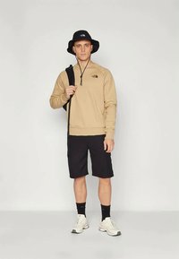 Μπεζ φλις φούτερ, μαύρη βερμούδα, άσπρα αθλητικά παπούτσια και μαύρο καπέλο μπόοτ, με λογότυπο στο φούτερ και το καπέλο. Κεντρική φιγούρα με σακίδιο .