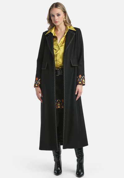 Dugi crni kaput s raznobojnim izvezenim detaljima na rukavima, šivenim detaljima i dva prednja džepa; uparen sa žutom satenskom košuljom.