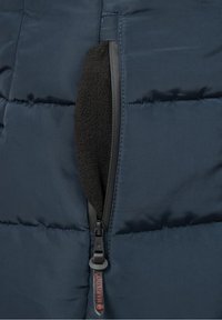 Mörkblå vadderad jacka med en ficka med dragkedja, utrustad med ett svart fleecefoder och texturerat tyg. Sömmen är synlig längs kanterna.