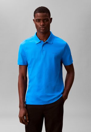 Bărbat purtând un tricou polo cu mânecă scurtă, de culoare albastru deschis, și pantaloni negri, stând în picioare pe un fundal gri simplu, cu fața spre înainte.