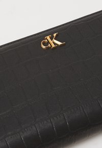 Melnā mākslīgā ādas maciņš ar krokodila ādas rakstu, ar zelta "CK" logo priekšpusē. Kompakts dizains ar gludu apdari.