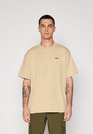 Beige oversized t-skjorte med rund hals, korte ermer og et subtilt teksturert mønster. Har en liten svart merkeetikett på brystet.