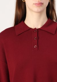 Burgundowy sweter polo o miękkiej fakturze, z klasycznym kołnierzykiem i trzema guzikami. Nowoczesny i minimalistyczny design.