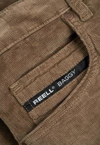 Un bolsillo de pantalones de pana marrón con una etiqueta negra visible que dice "REELL BAGGY" y un botón de cobre con el grabado "Reell".