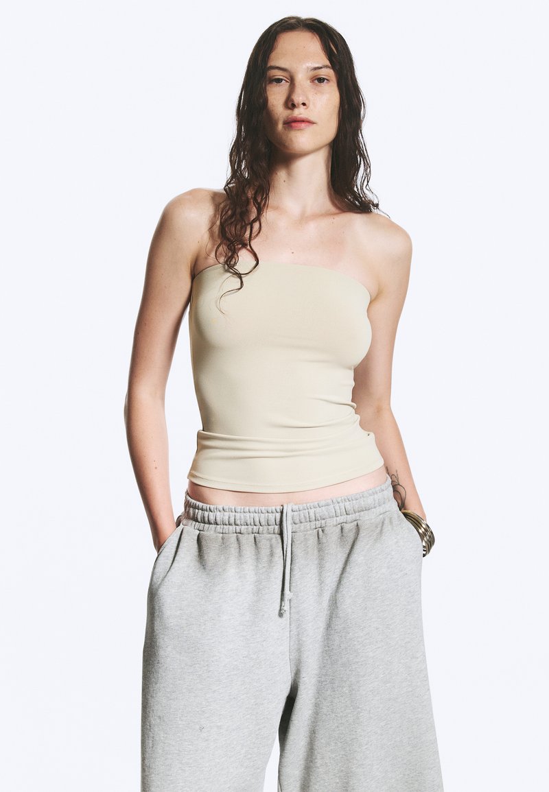 Femme aux longs cheveux foncés portant un haut beige sans bretelles et un pantalon de survêtement gris ample, les mains dans les poches, sur un fond uni.