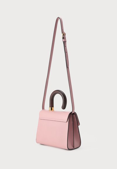Sac à main en cuir rose avec une poignée incurvée marron foncé et une longue bandoulière rose réglable, présenté sur un fond uni.