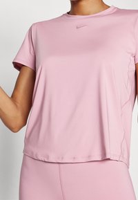 Světlá růžová sportovní tričko z hladké, lehké látky. Má kulatý výstřih, krátké rukávy a jemné logo Nike na hrudi.