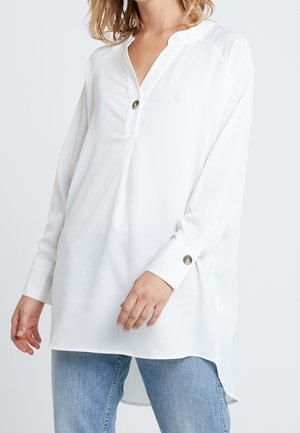 Femme portant un chemisier blanc ample à manches longues avec poignets et col boutonnés, associé à un jean en denim bleu clair.