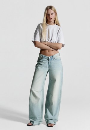 Ung kvinde med langt blondt hår iført en kort hvid T-shirt og vidde, falmede blå jeans, stående med armene krydsede.