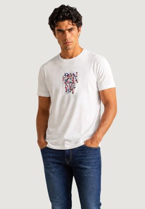 Homme aux cheveux bouclés et foncés portant un T-shirt blanc Armani Exchange avec un texte coloré et un jean bleu, debout les mains dans les poches.