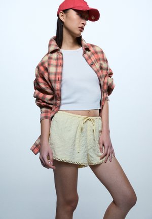 Noor naine kannab korallikarva ruudulist särki valge lühikese toppi peal, kollaseid pitsist lühikesi pükse ja punast pesapalli nokamütsi, poseerides heledal taustal.