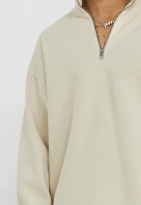 Beige mikina s čtvrtinovým zipem, volného střihu, s žebrovanou strukturou a metalickým zipem. Má spadlá ramena a žádné viditelné vzory.