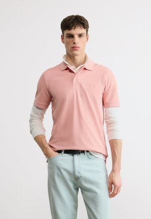 Νέος άνδρας που φοράει ροζ κοντομάνικο πόλο πάνω από λευκό μακρυμάνικο πουκάμισο και ανοιχτόχρωμο μπλε τζιν, στέκεται με το ένα χέρι στην τσέπη.