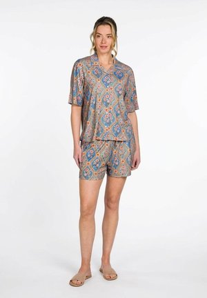 Woman standing wearing a matching blue and orange floral patterned short-sleeve shirt and shorts set with beige slide sandals.