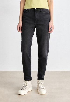 Osoba ubrana w czarne, prostokątne jeansy z podwyższonym stanem oraz kremowe botki na sznurowadła, stojąca na jasnej podłodze na tle jednolitej ściany.