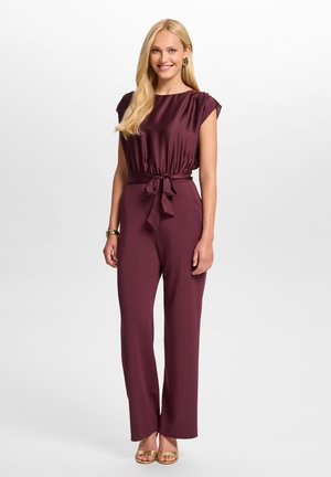 Femme aux longs cheveux blonds portant une combinaison sans manches violet foncé avec une ceinture nouée à la taille et des sandales dorées, souriant à la caméra.