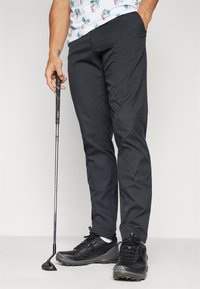 Černé golfové kalhoty s hladkou texturou a štíhlým střihem, doplněné černými sportovními botami a golfovou holí, držena uvolněně v ruce.