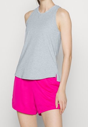 Kvinde iført ærmeløs lysegrå top og lyserøde shorts, stående mod en ensfarvet lys baggrund.