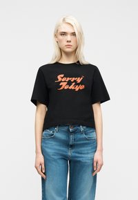 Νεαρό άτομο με μακριά ξανθά μαλλιά, που φοράει μια μαύρη cropped μπλούζα με πορτοκαλί κείμενο "Sorry Tokyo" και μπλε τζιν, στέκεται μπροστά σε ένα λευκό φόντο.