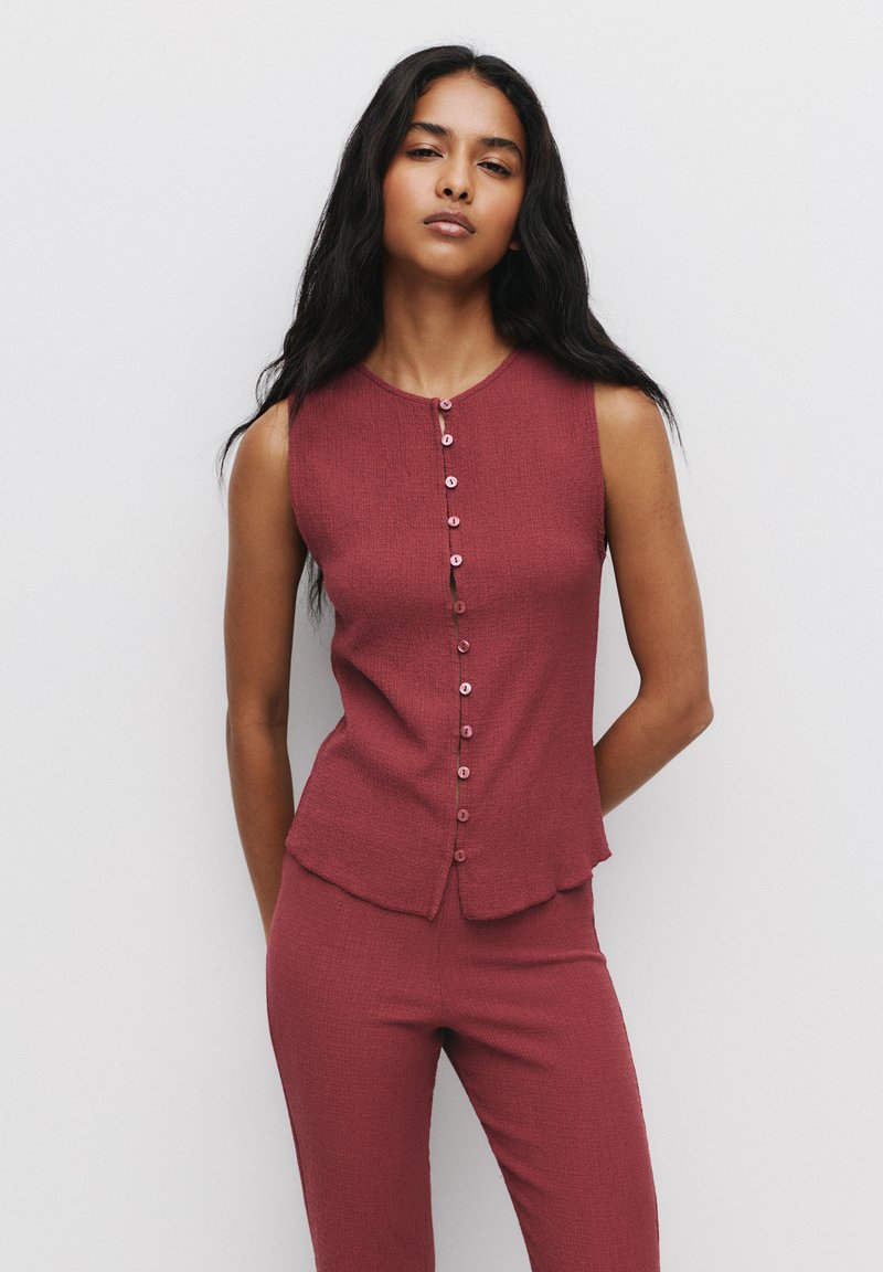 Maroon texturerad ärmlös topp med rund halsringning och knappar framtill; kombinerad med matchande åtsittande byxor. Slät material, avslappnad design.