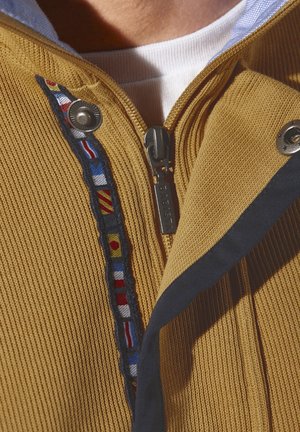 Gelbe Zip-Jacke mit Rippstruktur, ausgestattet mit einem marineblauen Besatz und einem mehrfarbigen Bändchenakzent entlang des Reißverschlusses. Sichtbarer silberner Reißverschluss-Zug.