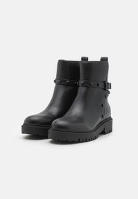 Botas de tobillo de cuero negro con suela gruesa, que cuentan con una correa adornada con tachuelas cuadradas. Textura suave y tira de agarre para ponerlas fácilmente.